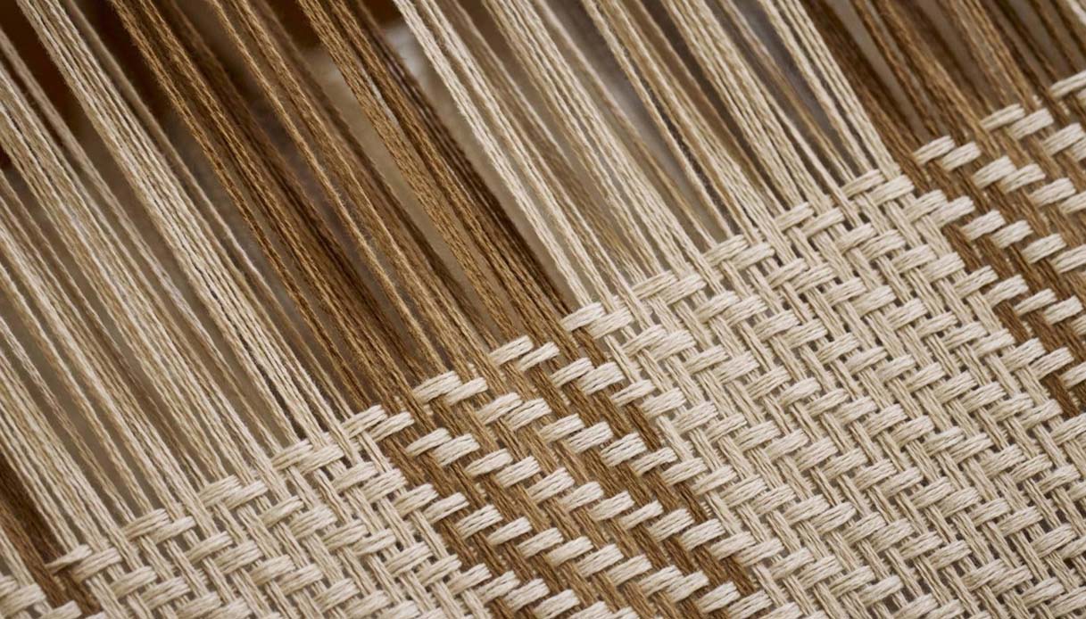 Macro di tessuto in fibra beige lavorato a telaio manuale con ordito in evidenza e intreccio a punto tela, dettaglio mostra Loro Piana Milano Design Week 2026
