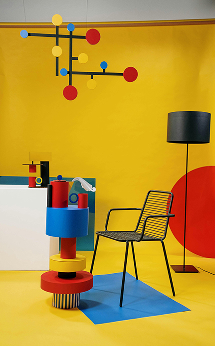 Allestimento ispirato al Memphis Group con parete gialla, oggetti cilindrici colorati impilati, mobile a struttura nera e lampadario geometrico sospeso