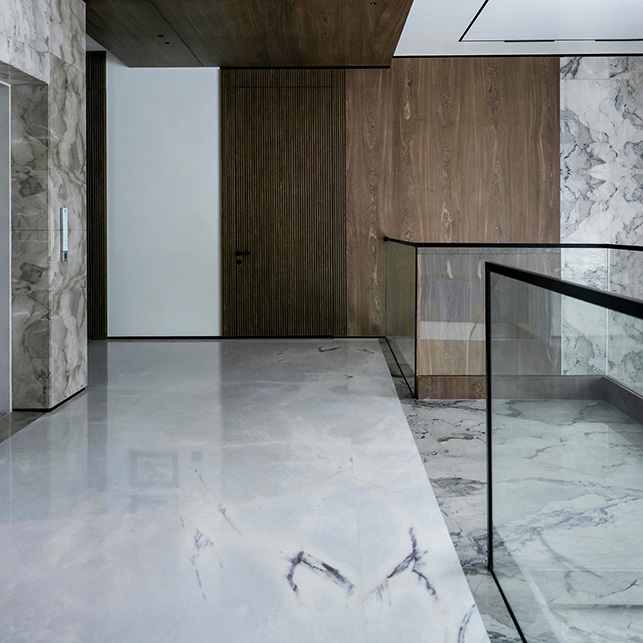 Ingresso con pavimento in marmo bianco con venature grigie a grande formato, parete con pannelli in legno a doghe verticali e ringhiera in vetro e metallo nero