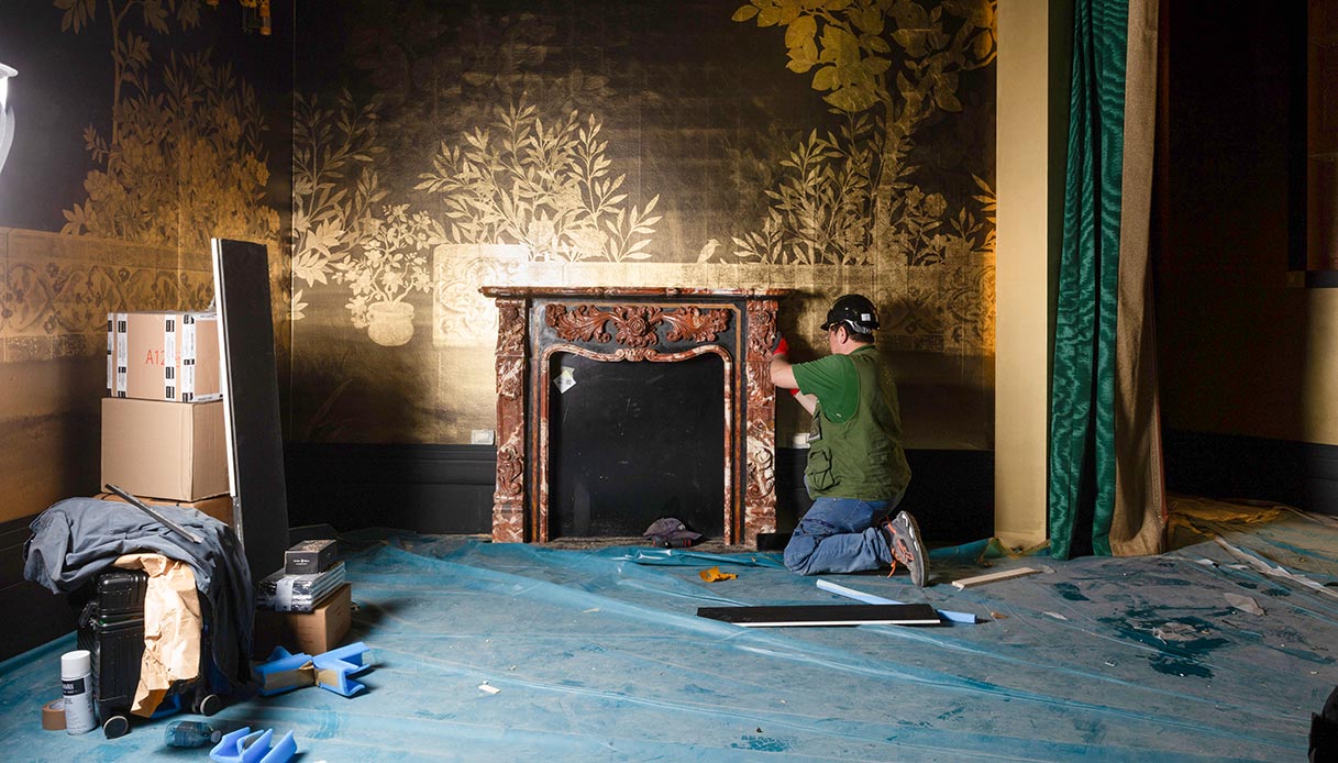 Operaio al lavoro sull'allestimento di Aurea an Architectural Fiction con camino in marmo scolpito, carta da parati dorata con motivi floreali e teli di protezione sul pavimento