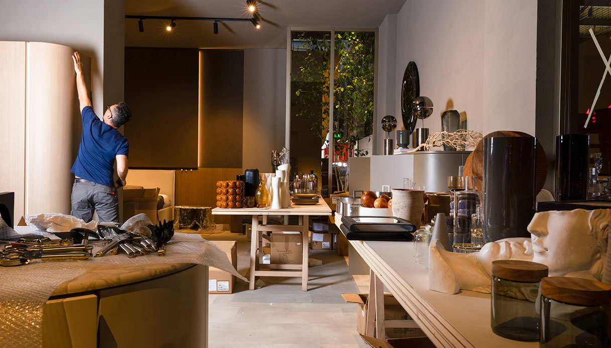 Operaio al lavoro nell'allestimento di uno stand di EuroCucina con oggetti di design, complementi d'arredo e imballaggi ancora aperti sul tavolo