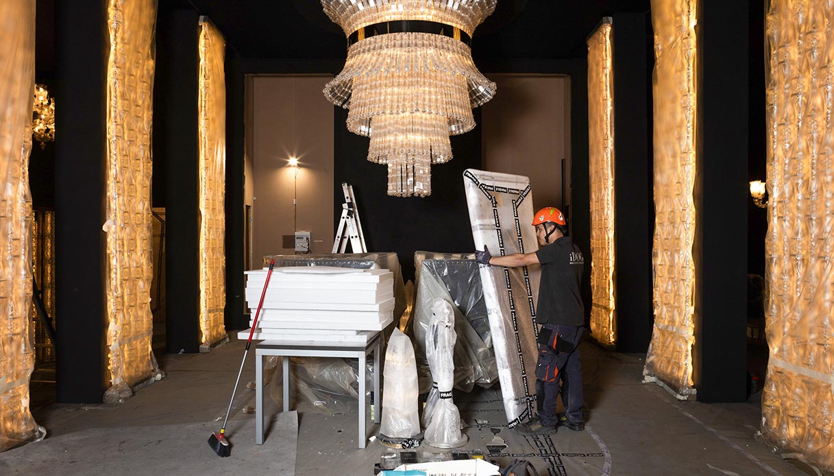 Operaio con casco arancione al lavoro nell'allestimento di uno stand con lampadario in cristallo e colonne dorate durante il making of del Salone del Mobile 2026