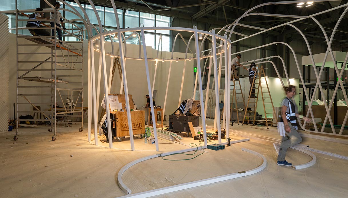 Maestranze al lavoro sull'allestimento di un padiglione del Salone del Mobile 2026 con strutture circolari in metallo bianco, scale e impalcature