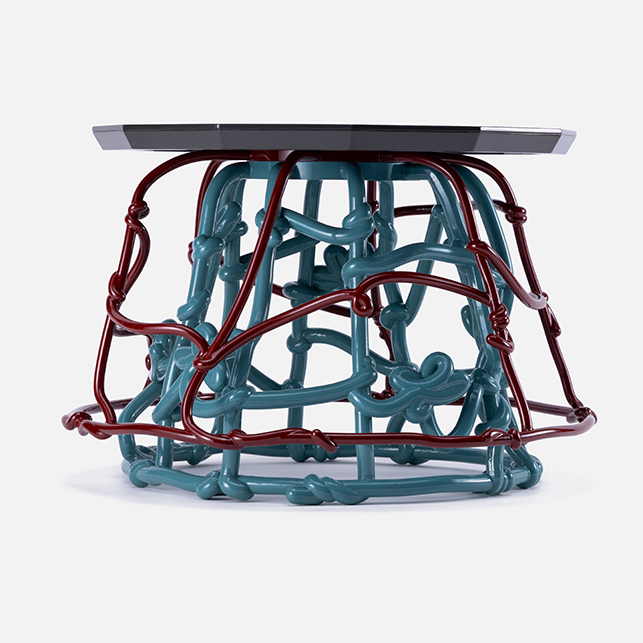 Soban con piano grigio scuro e base in intreccio di tubature smaltate petrolio e bordeaux su fondo bianco neutro