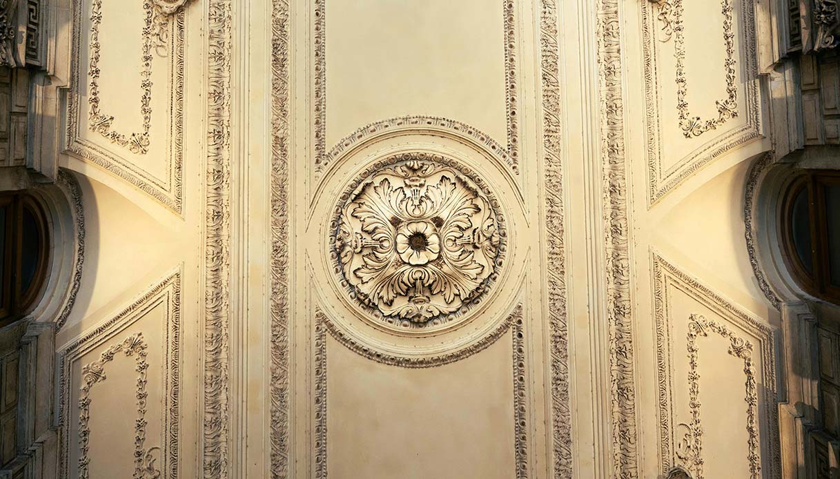 Soffitto con elaborate modanature in stucco bianco avorio, rosone centrale a motivo floreale e pannelli decorativi in stile rococ&ograve;