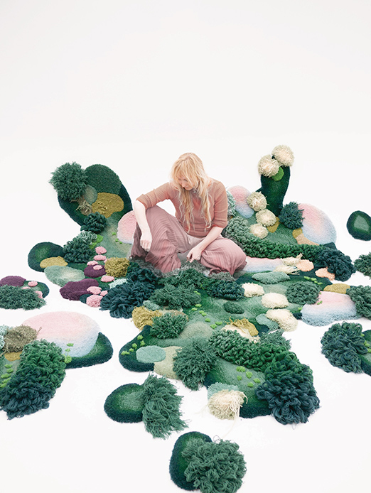 Artista svedese Alfhild K&uuml;lper seduta tra elementi scultorei in lana a forma di cespugli e prati in verde muschio, rosa e giallo paglierino su pavimento bianco