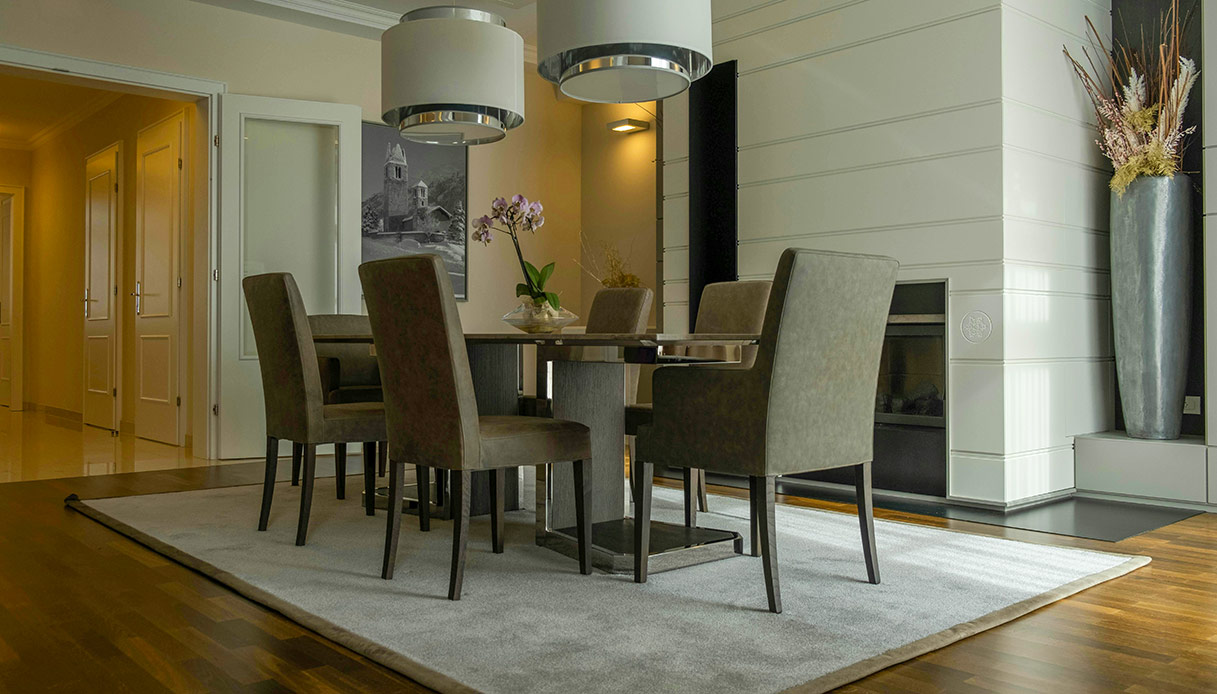 Sala da pranzo con tavolo rotondo, sei sedie imbottite grigio tortora e tappeto chiaro su parquet in legno scuro, con lampadari a sospensione cromati