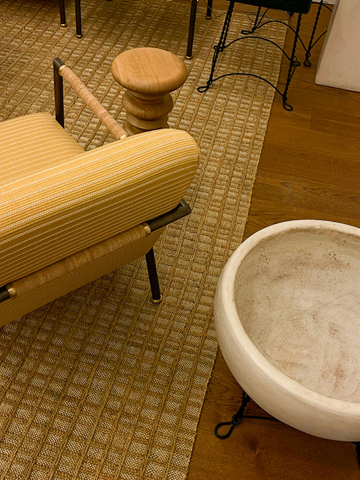 Dettaglio di tappeto in fibra naturale intrecciata posato su parquet in legno chiaro, con divano giallo a righe e sgabello in legno