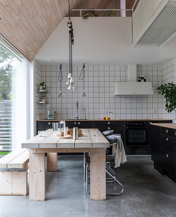 Cucina in stile nordico con tavolo rettangolare in legno grezzo, panche abbinate, ante nere, piano di lavoro in legno, piastrelle bianche a griglia e pavimento in cemento levigato