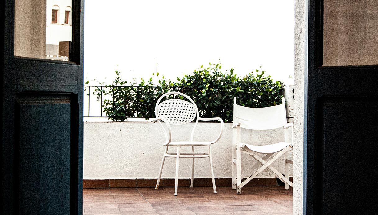 Terrazza con pavimento in cotto, due sedie bianche in metallo e legno, grande vaso con siepe verde, vista dall'interno attraverso una porta aperta