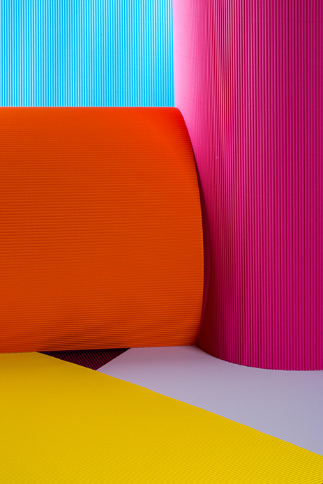 Composizione astratta con superfici ondulate in cartone colorato nei toni arancione, fucsia, azzurro e giallo, con texture a righe fini in primo piano