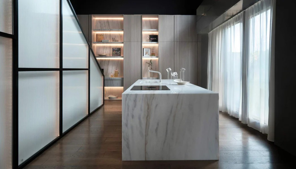 Cucina TM Italia Avignon design Sabatini con isola monolitica interamente rivestita in marmo Calacatta bianco, colonne attrezzate con illuminazione calda e parete divisoria in vetro acanalato