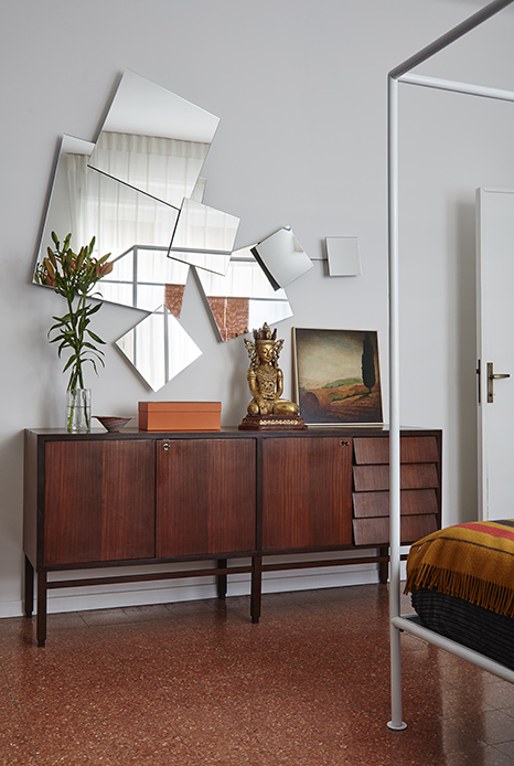 Cassettone in teak con ante e cassetti, sopra Buddha dorato su base, scatola Herm&egrave;s arancio, dipinto paesaggistico incorniciato e vaso con fiori, specchiera con frammenti di specchio irregolari alla parete, struttura letto a baldacchino a destra