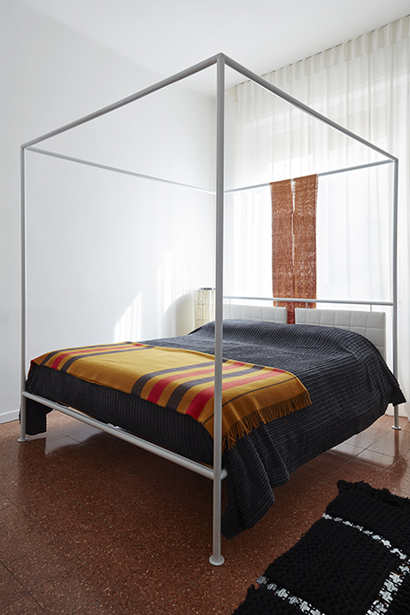 Camera da letto con letto a baldacchino in tubolare metallico grigio, coperte a righe giallo ocra rosso e grigio, pavimento in graniglia rossa, tende bianche alla finestra e tessuto africano arancione appeso