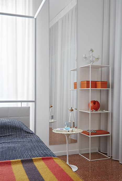 Camera da letto con specchiera componibile in frammenti metallici su parete bianca, scaffale bianco con scatole arancio Herm&egrave;s e oggetti decorativi, tavolino rotondo Saarinen in marmo con caraffa in vetro, letto sullo sfondo
