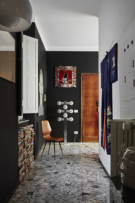 Corridoio lungo con pareti nere, lampada da terra a stelo con sfere in vetro bianco, sedia in legno curvato, kimono blu con decorazioni appeso a destra, quadro con tenda rossa sul muro e pile di libri a sinistra