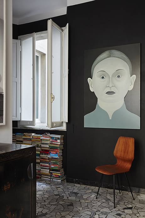 Corridoio con pareti nere e pavimento in marmo irregolare, grande dipinto raffigurante volto frontale di donna in toni grigi e verdi, sedia in legno curvato rossiccio con gambe nere, pile di libri a sinistra