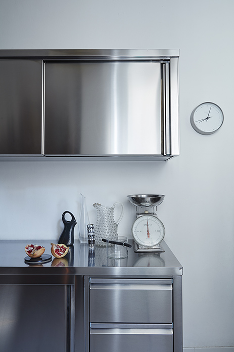 Piano di lavoro in acciaio inox con bilancia da cucina vintage, caraffe in vetro soffiato trasparente, melograno tagliato, pensile superiore in acciaio lucido con ante scorrevoli e orologio rotondo a parete