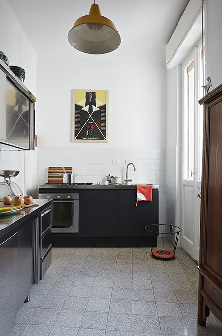 Cucina stretta con moduli in acciaio inox nero opaco, piano in acciaio, lampada a campana giallo ocra sul soffitto, poster grafico di Vico Magistretti alla parete, armadio in legno scuro a destra, finestra con luce naturale