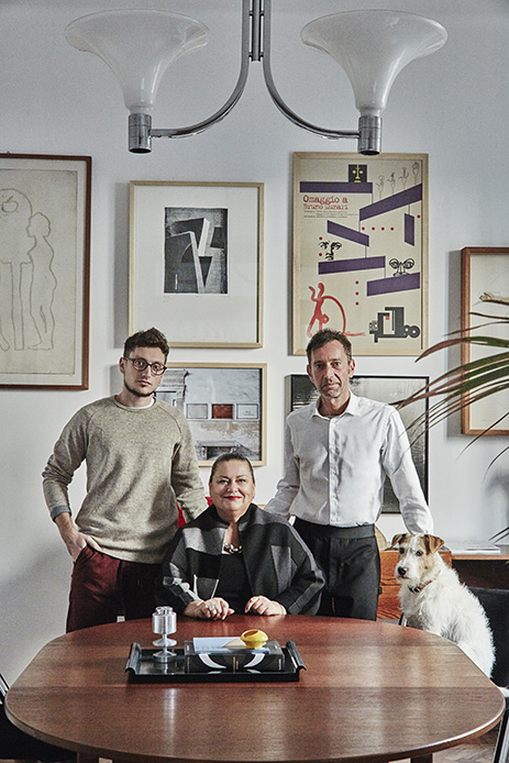 Tre persone attorno al tavolo rotondo in legno nella sala da pranzo, lampadario a due coppe bianche sopra, parete con galleria di quadri e poster, fox terrier bianco accanto all'uomo a destra