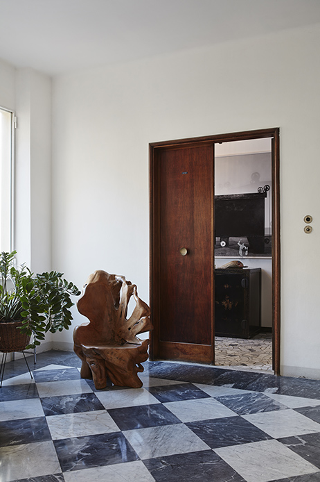 Ingresso con pavimento a scacchiera in marmo grigio e bianco, sedia scultorea in legno di ulivo grezzo, porta in legno scuro aperta sulla cucina, pianta verde in vaso a sinistra