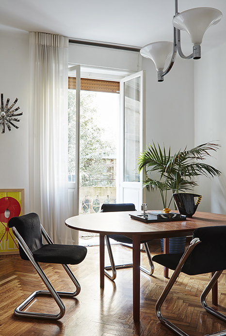 Sala da pranzo luminosa con tavolo rotondo in legno, sedie a sbalzo in tubolare cromato con seduta nera, lampadario a due coppe bianche in acciaio cromato, finestra aperta sul verde, palma in vaso a destra