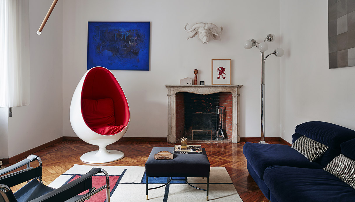 Soggiorno con Ball Chair bianca con interno rosso, divano in velluto blu notte, camino in marmo con decorazioni sul mantello, dipinto blu su parete sinistra e scultura testa di bufalo bianca in alto