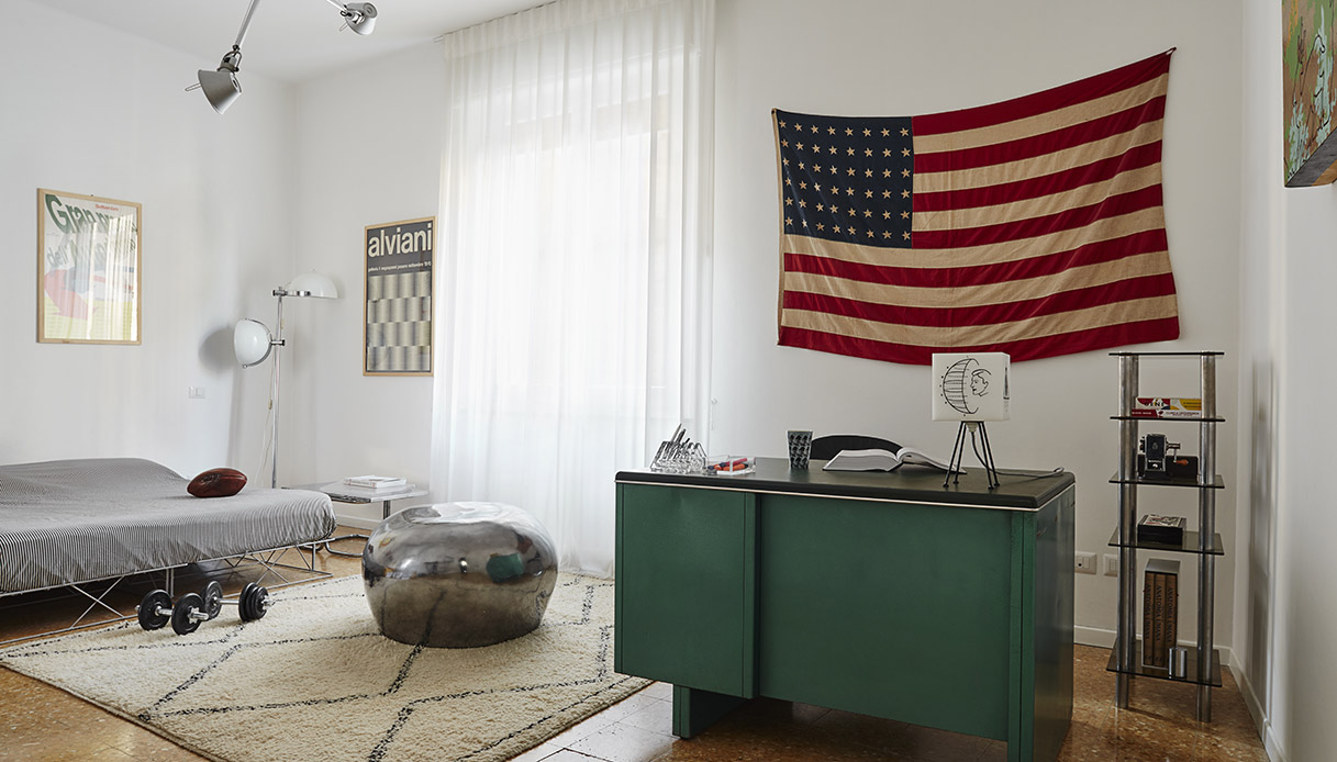 Camera da letto con scrivania vintage verde scuro, tappeto berbero bianco con motivi geometrici neri, grande bandiera americana d'epoca appesa alla parete, lampada da terra cromata con globo bianco e poster Alviani