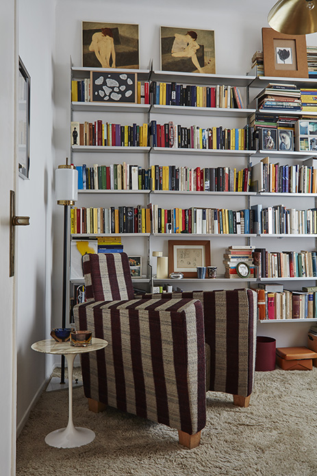 Angolo studio con poltrona a righe verticali bordeaux e beige, sistema di scaffali in acciaio a parete pieno di libri colorati e opere d'arte, tavolino rotondo Saarinen in marmo, lampada da terra con paralume bianco