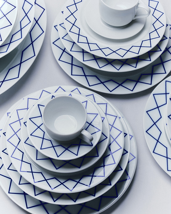 Servizio di piatti in porcellana bianca con decorazione geometrica blu a stelle e rombi sovrapposti in pile su superficie bianca, tazzine da caff&egrave; bianche sopra ogni set