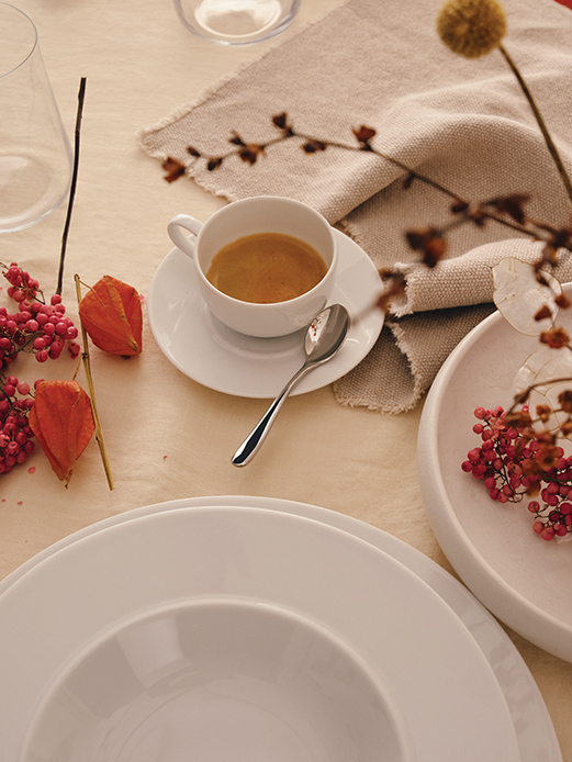 Apparecchiatura con piatti fondi e piani in porcellana bianca, tazzina da caff&egrave; con caff&egrave; espresso e cucchiaino in acciaio, fiori secchi arancio e bacche rosa su tovaglia beige e lino grezzo
