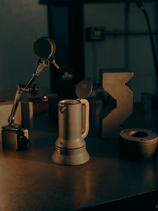 Caffettiera 9090 di Richard Sapper per Alessi in finitura metallica nera opaca con coperchio brandizzato C.P. Company su piano di lavoro industriale in acciaio, con strumenti di misura sullo sfondo