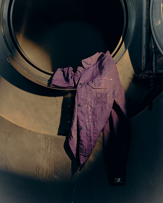 Giacca viola con patch C.P. Company x Alessi appesa all'apertura di un grande macchinario industriale cilindrico in acciaio ossidato, con gocce di colore visibili sul tessuto