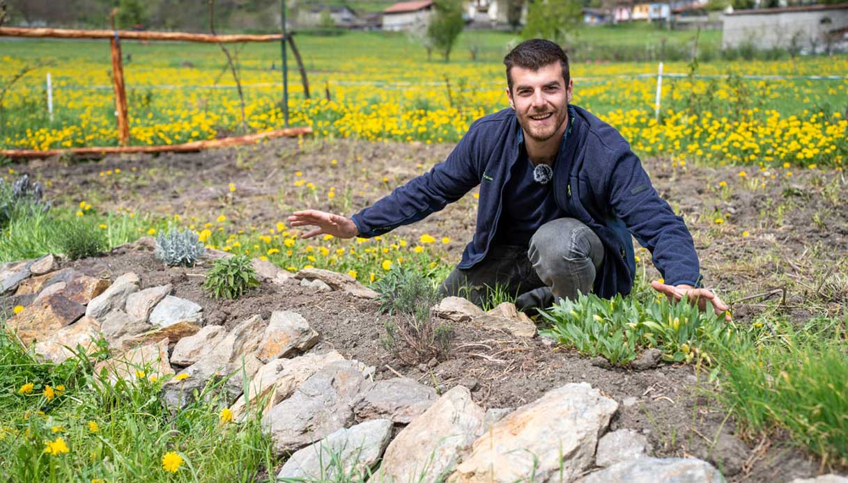 Gian Marco Mapelli accovacciato in un orto con terreno lavorato e bordura in pietra, piante aromatiche appena trapiantate e prato fiorito sullo sfondo