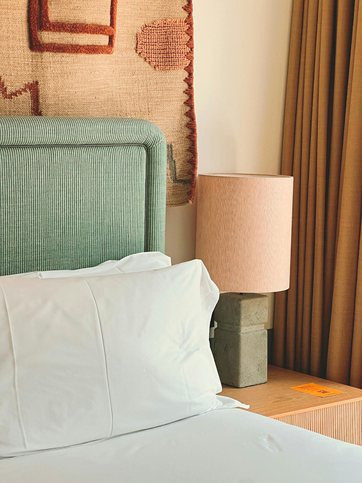 Dettaglio camera da letto con testiera imbottita verde a coste, lampada da comodino con base in cemento e paralume rosa, arazzo in lana con motivo geometrico terracotta