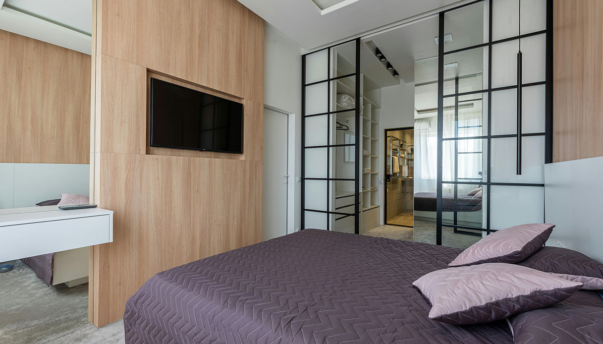 Camera da letto con pannello in legno chiaro che integra una TV a schermo piatto, letto con copriletto viola e parete divisoria in vetro e acciaio nero