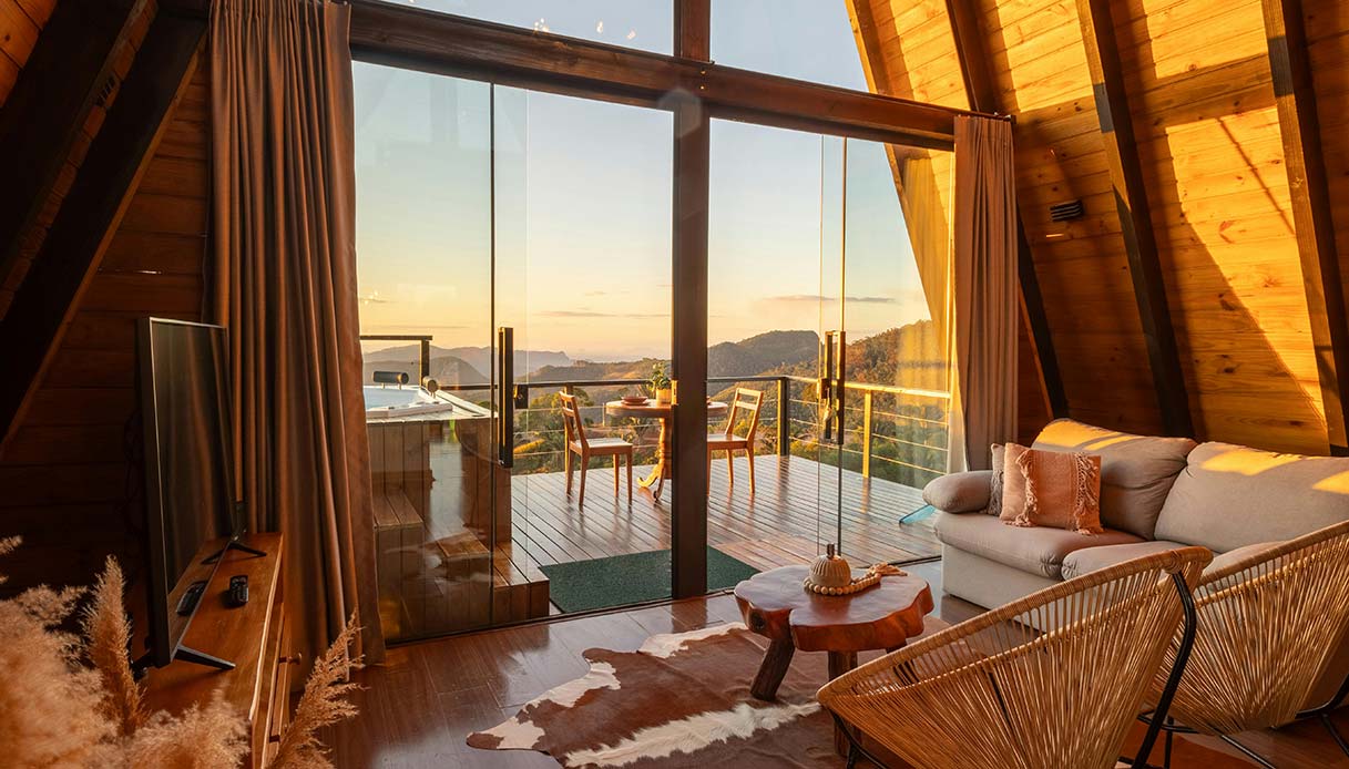 Interno di chalet a forma di A con vetrata aperta su terrazza con vista su montagne al tramonto, divano beige, tavolino in legno nodoso e poltrona in corda intrecciata