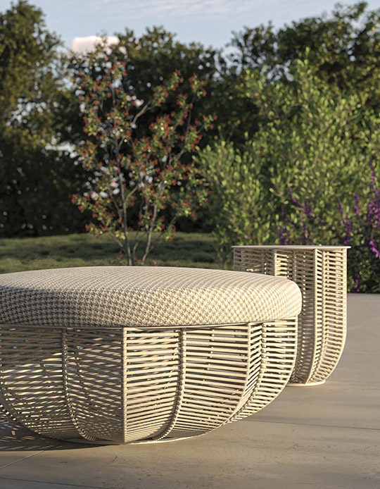 Due pouf outdoor con intreccio perimetrale in corda beige e cuscino in tessuto pied-de-poule crema su pavimento in pietra, con vegetazione sullo sfondo