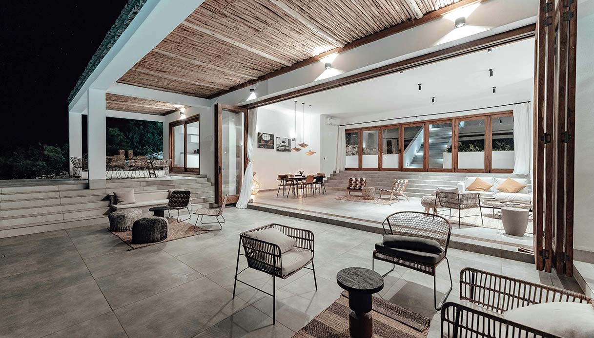 Villa fotografata di notte con spazio living aperto su terrazza, sedie in corda intrecciata, soffitto a canne, pareti bianche e illuminazione puntuale