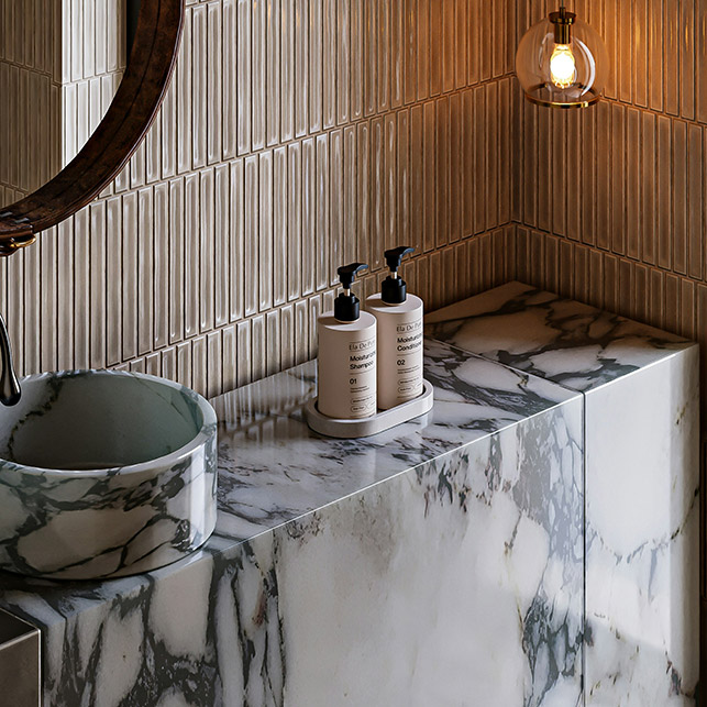 Dettaglio bagno con parete rivestita in piastrelle rettangolari verticali beige con texture costolata e fuga tono su tono, lavabo a catino tondo in marmo verde, piano in marmo bianco con venature grigie, lampada a sospensione in vetro ambra