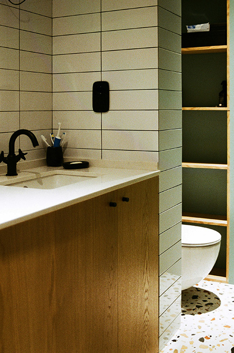 Bagno con piastrelle rettangolari in posa verticale verde salvia con fughe grigio scuro, mobile sottolavabo in rovere chiaro con piano bianco, rubinetteria nera, pavimento in terrazzo e nicchia con mensole a vista in legno