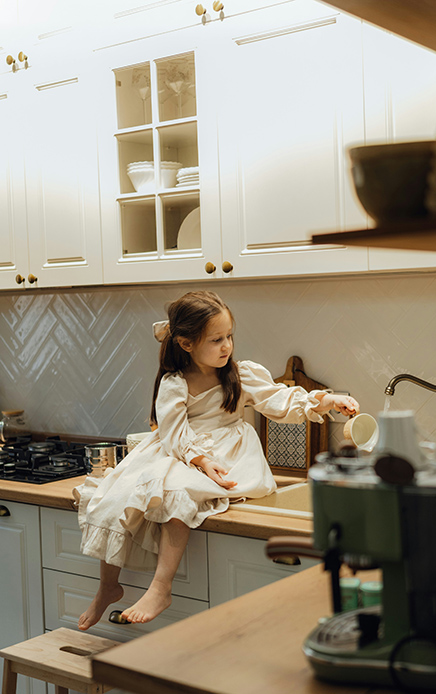 Bambina in abito bianco seduta sul piano di una cucina classica bianca, intenta a versare qualcosa in una tazza accanto alla macchinetta del caffè