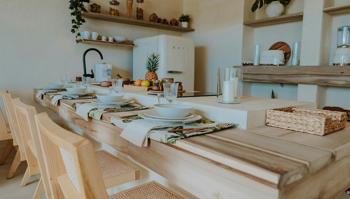 Cucina e sala da pranzo con tavolo in legno grezzo apparecchiato, sedie in rattan, frigorifero Smeg bianco e mensole in legno con barattoli