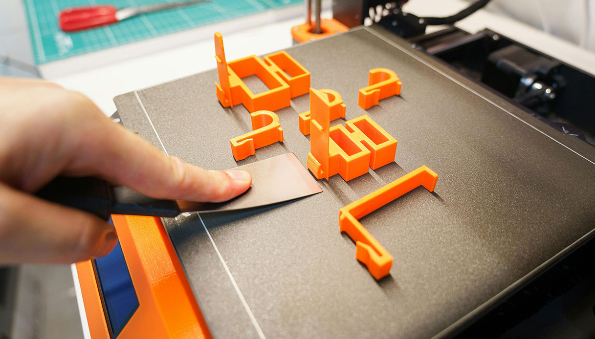 Mano che rimuove con spatola componenti stampati in 3D color arancione dal piano di una stampante FDM