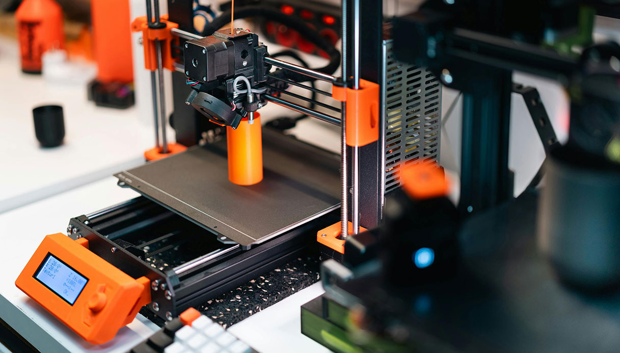 Stampante 3D FDM con struttura arancione e nera, testina in movimento sul piano di stampa e schermo LCD con parametri