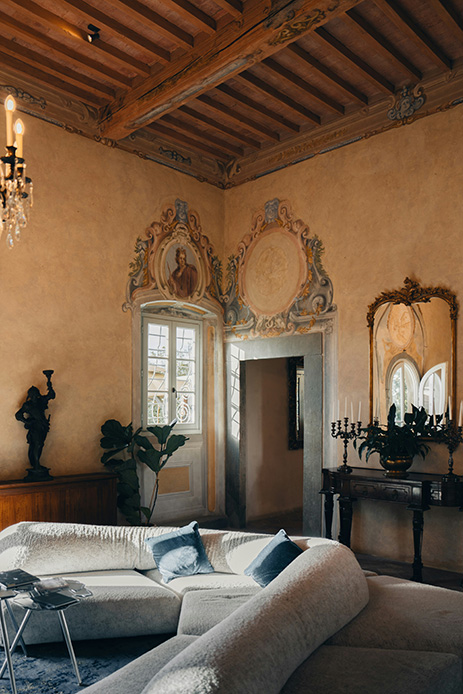 Sala con travi in legno a vista, affreschi con cornici barocche, divano in velluto grigio con cuscini blu, candelabri in ferro nero e specchio dorato