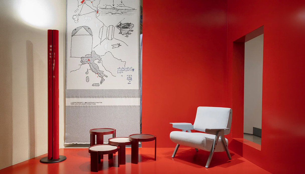 Quattro tavolini rotondi impilabili in lacca rossa bordeaux con piani in formica bianca, esposti su pavimento rosso con pannelli laccati rosso alle pareti, poltrona bianca con gambe inclinate a destra e arazzo con disegni geografici sullo sfondo
