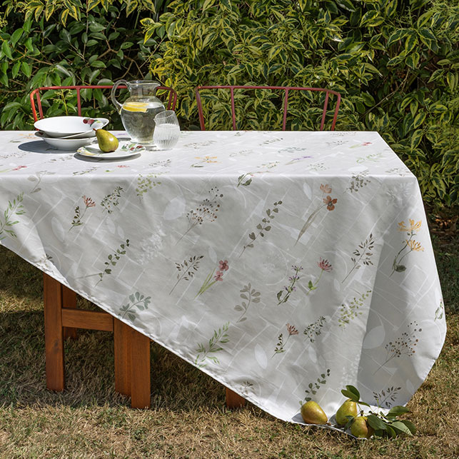 Tavolo da pranzo outdoor in legno con tovaglia bianca a motivo di fiori e erbe selvatiche in cotone lino e canapa, piatti bianchi, caraffa in vetro e pere, sedie in metallo rosso, giardino verde sullo sfondo