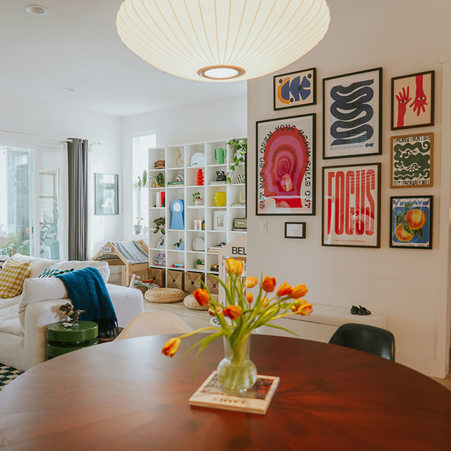 Soggiorno luminoso con gallery wall di poster grafici colorati, libreria bianca, divano bianco e tulipani arancioni sul tavolo