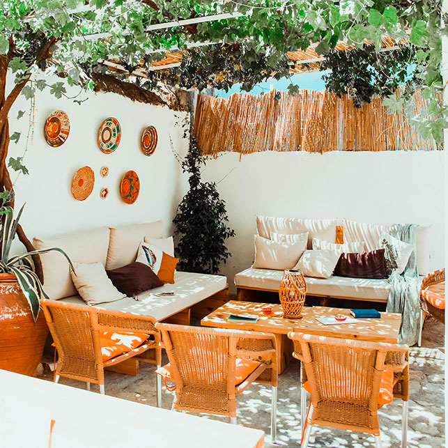 Spazio outdoor con sedie in vimini arancio, divani bassi con cuscini beige e grigi, tavolini in legno, piatti decorativi colorati alle pareti bianche, soffitto naturale di foglie di vite e separ&eacute; in cannucce
